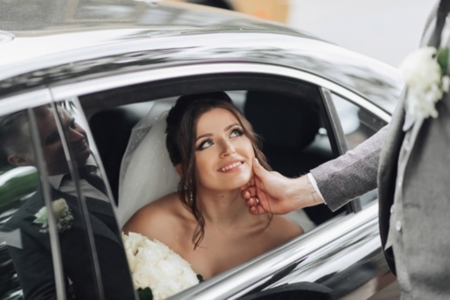Wedding photo. The bride sits in a Black Car Service with a smile, the groom gently holds the bride's chin. View through the car window. Beautiful makeup and hair. A luxurious white dress